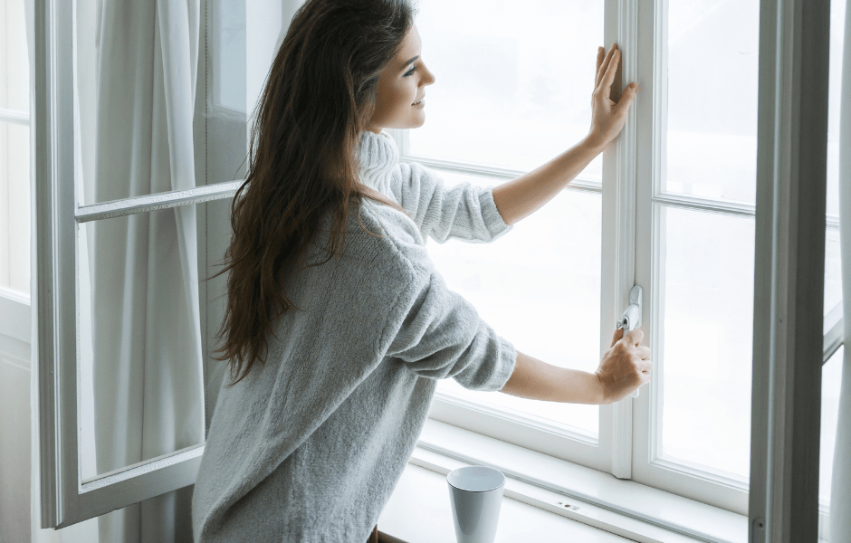 Chica abriendo la ventana practicable de su casa para que entre la luz y la luminosidad en su casa mientras toma su café